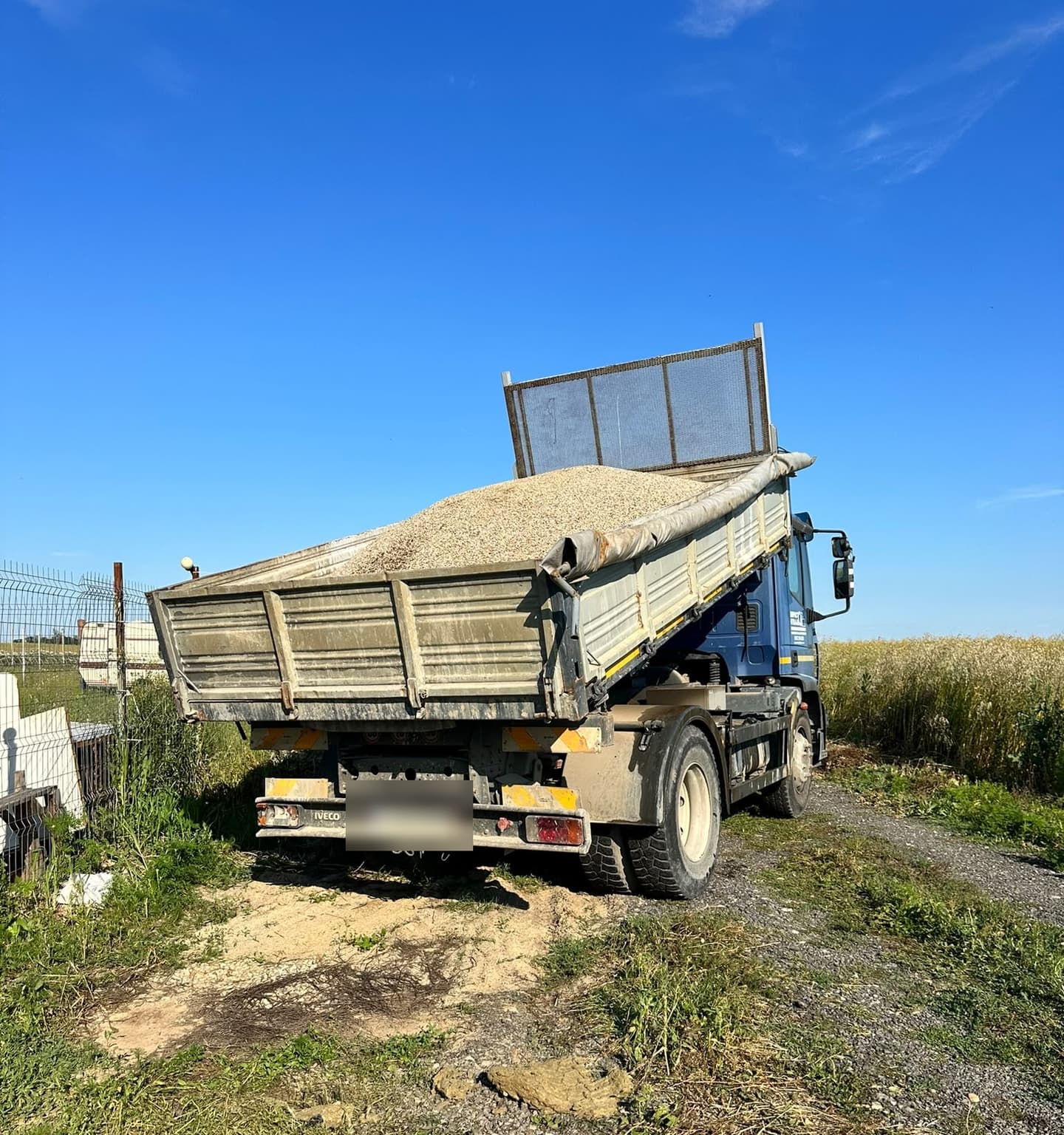 Basculantă Iveco cu bena ridicată descărcând balast pe un teren, lângă un câmp cultivat