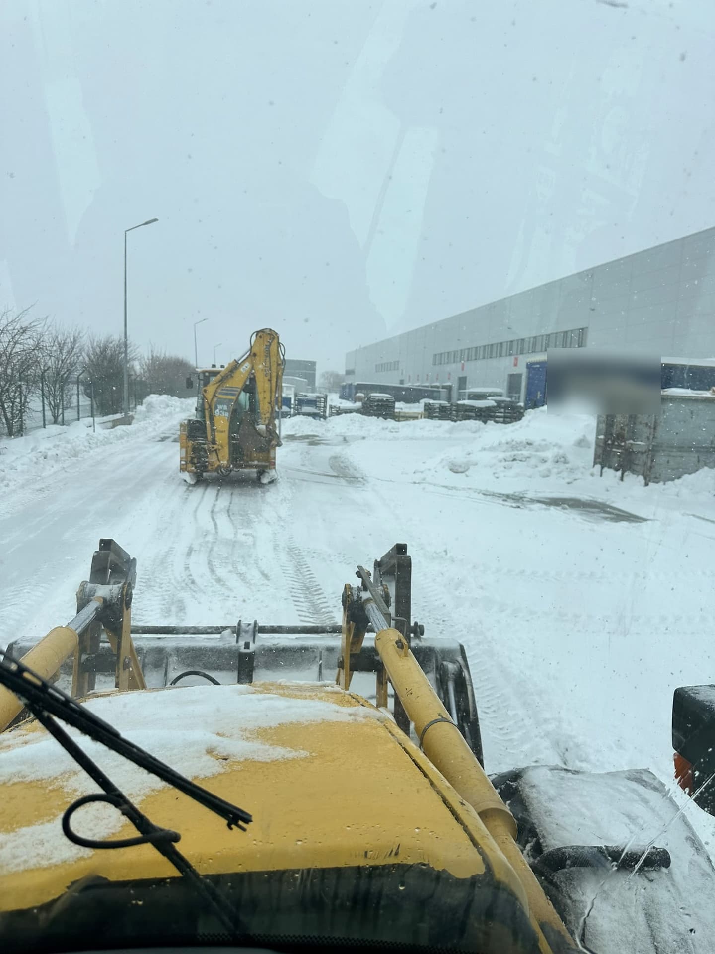 Buldoexcavator Komatsu WB93R curăță zăpada pe aleea unui parc logistic, văzut din cabina altui utilaj