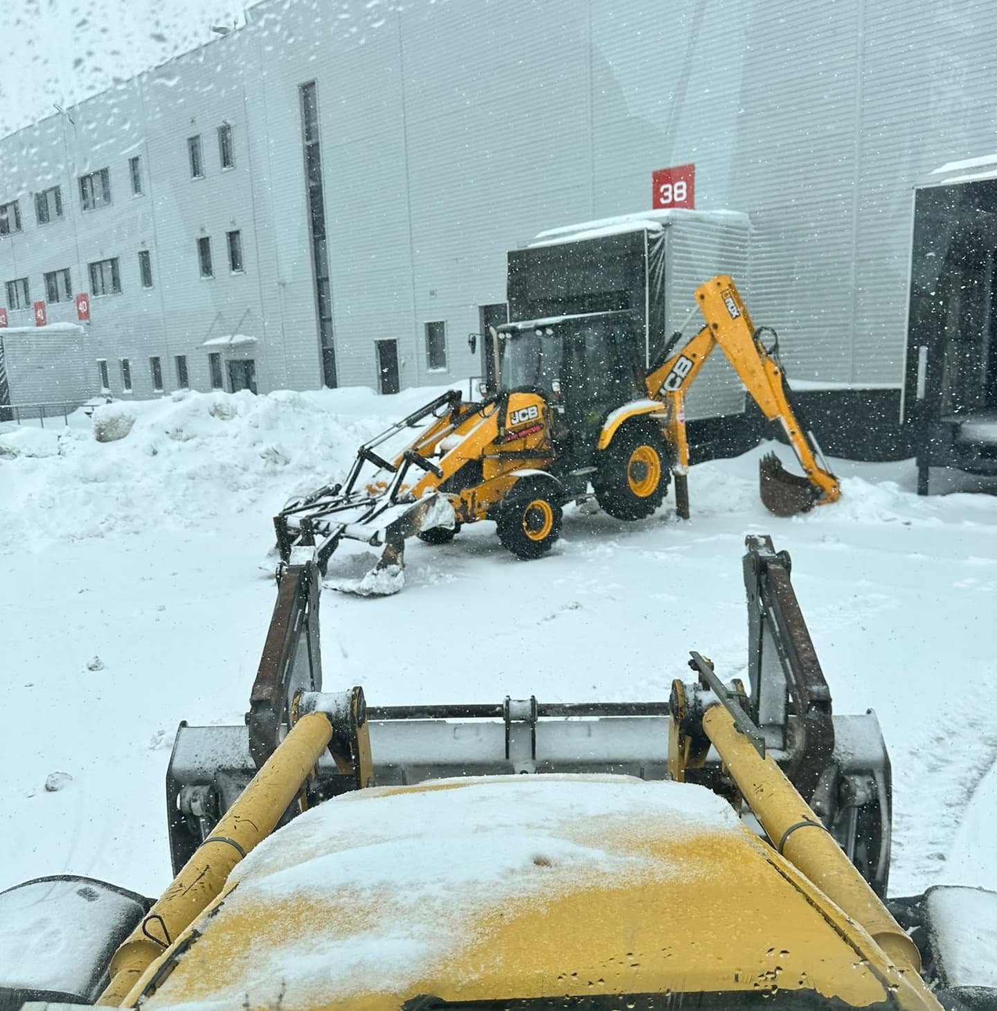 Buldoexcavator JCB deszăpezește platforma unui depozit logistic, văzut din cabina altui buldoexcavator