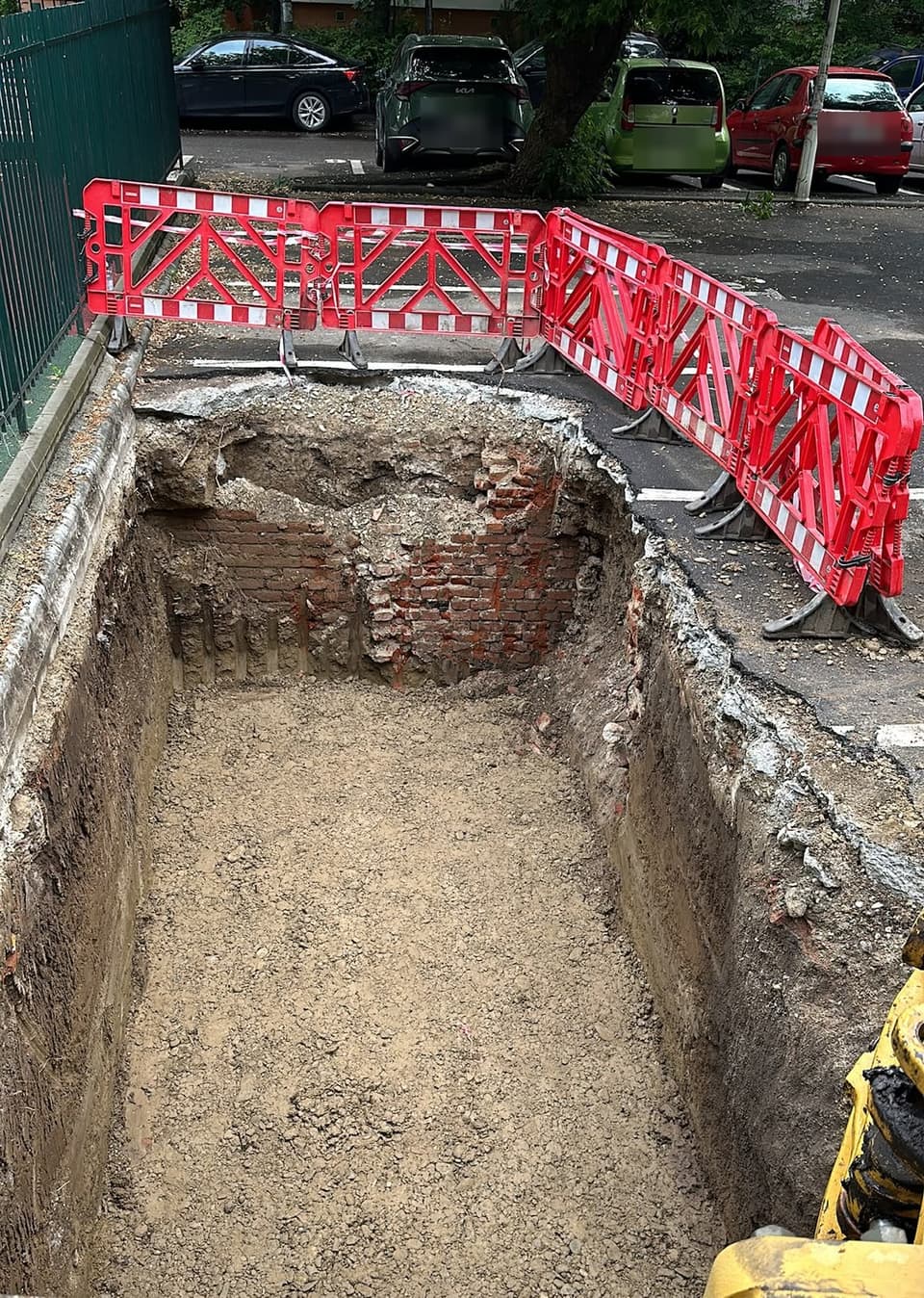 Groapă dreptunghiulară cu pereți drepți, excavată în asfaltul unei parcări de bloc și împrejmuită cu bariere