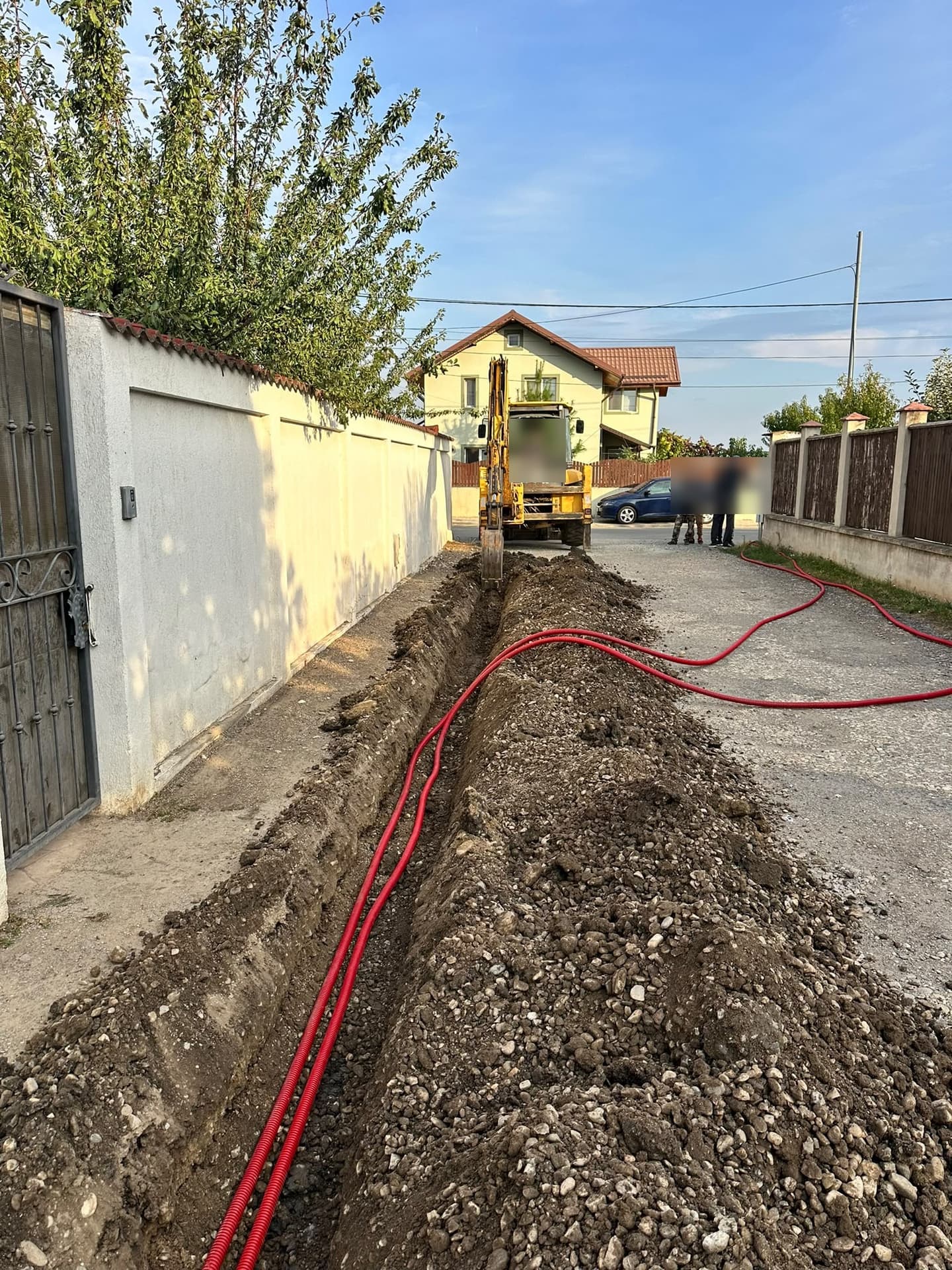 Buldoexcavator Komatsu sapă un șanț pe o stradă cu case; tuburi corugate roșii pentru cabluri pe margine